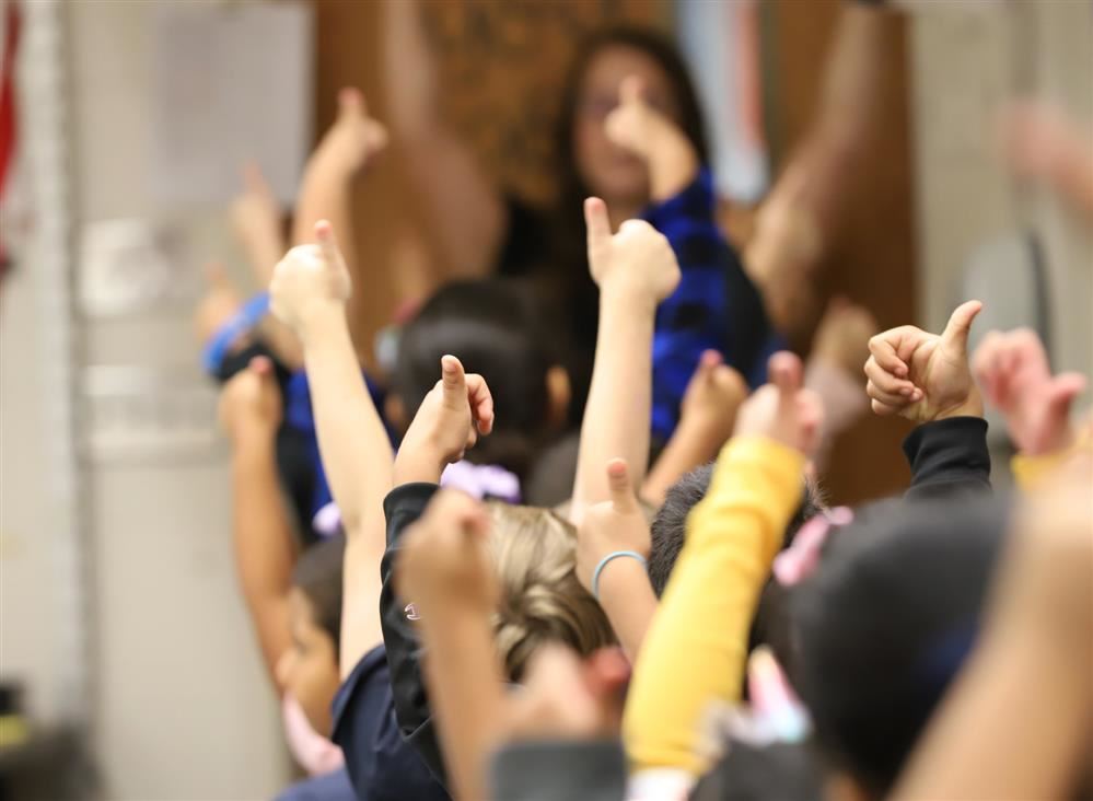 Students holding up thumbs-up with teacher standing in front of the classroom. 